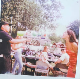 Backyard summer gathering with people at folding chairs and tables as a man playfully fires a large green water gun at a laughing woman in a red dress, trees and houses in the background.