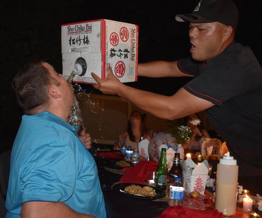 Man pouring sake from a boxed dispenser into a friend’s mouth at an outdoor nighttime party, table full of takeout containers, bottles, plates and candles.