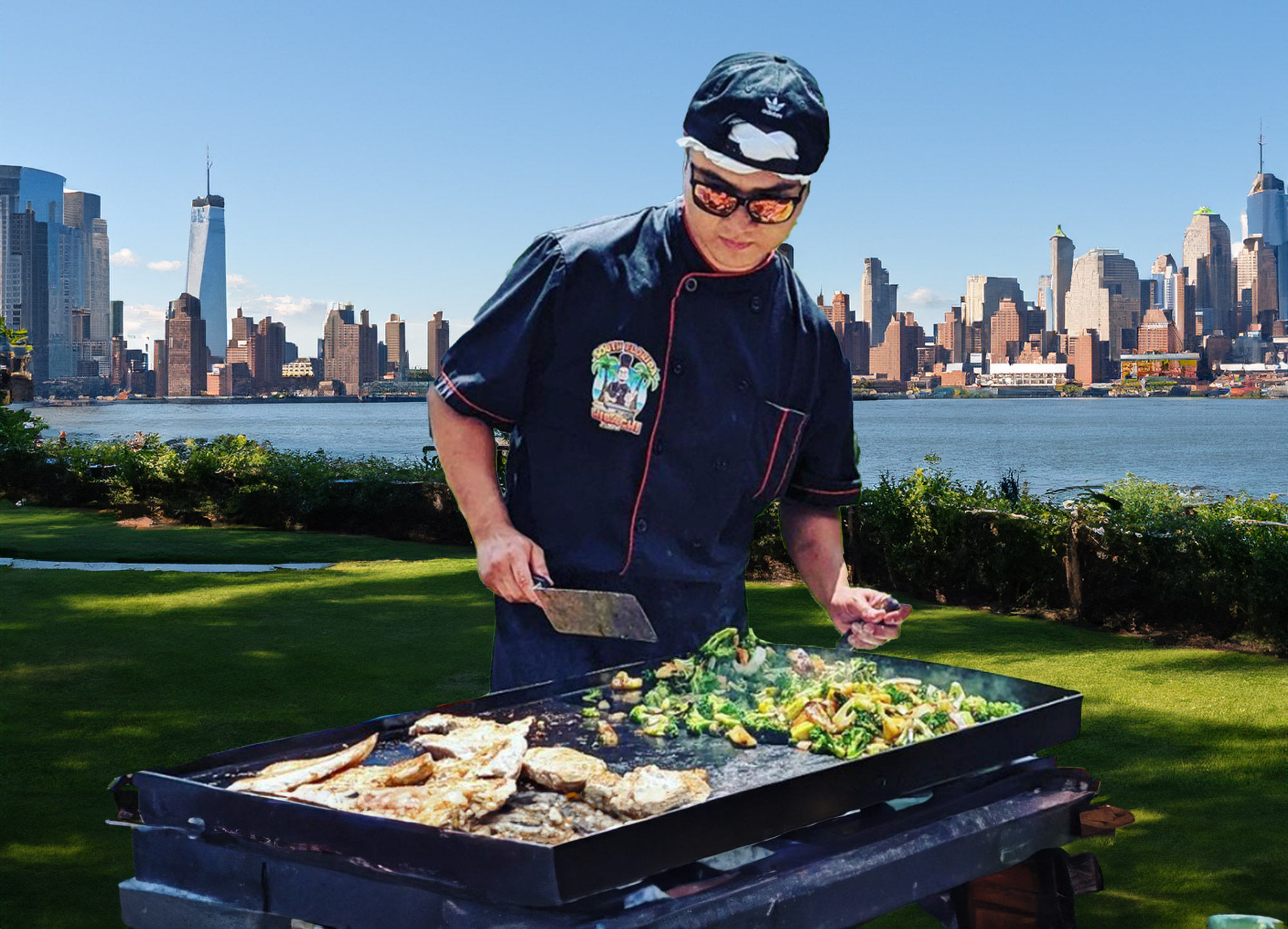 Chef grilling fish and sautéed vegetables on a flat-top griddle in a riverside park with the Manhattan skyline across the Hudson River in the background.