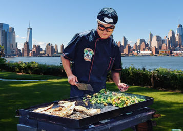 Chef grilling fish and sautéed vegetables on a flat-top griddle in a riverside park with the Manhattan skyline across the Hudson River in the background.