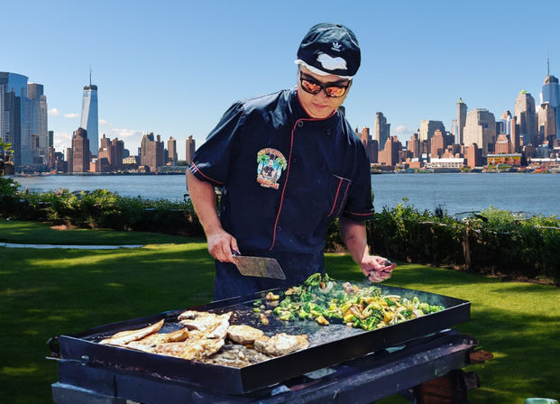 Chef grilling fish and sautéed vegetables on a flat-top griddle in a riverside park with the Manhattan skyline across the Hudson River in the background.