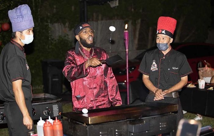 Nighttime outdoor hibachi scene with three chefs at a flat-top griddle — one in a red satin jacket theatrically flipping an egg with a spatula while two masked chefs in tall hats watch, condiment bottles and a tiki torch nearby.