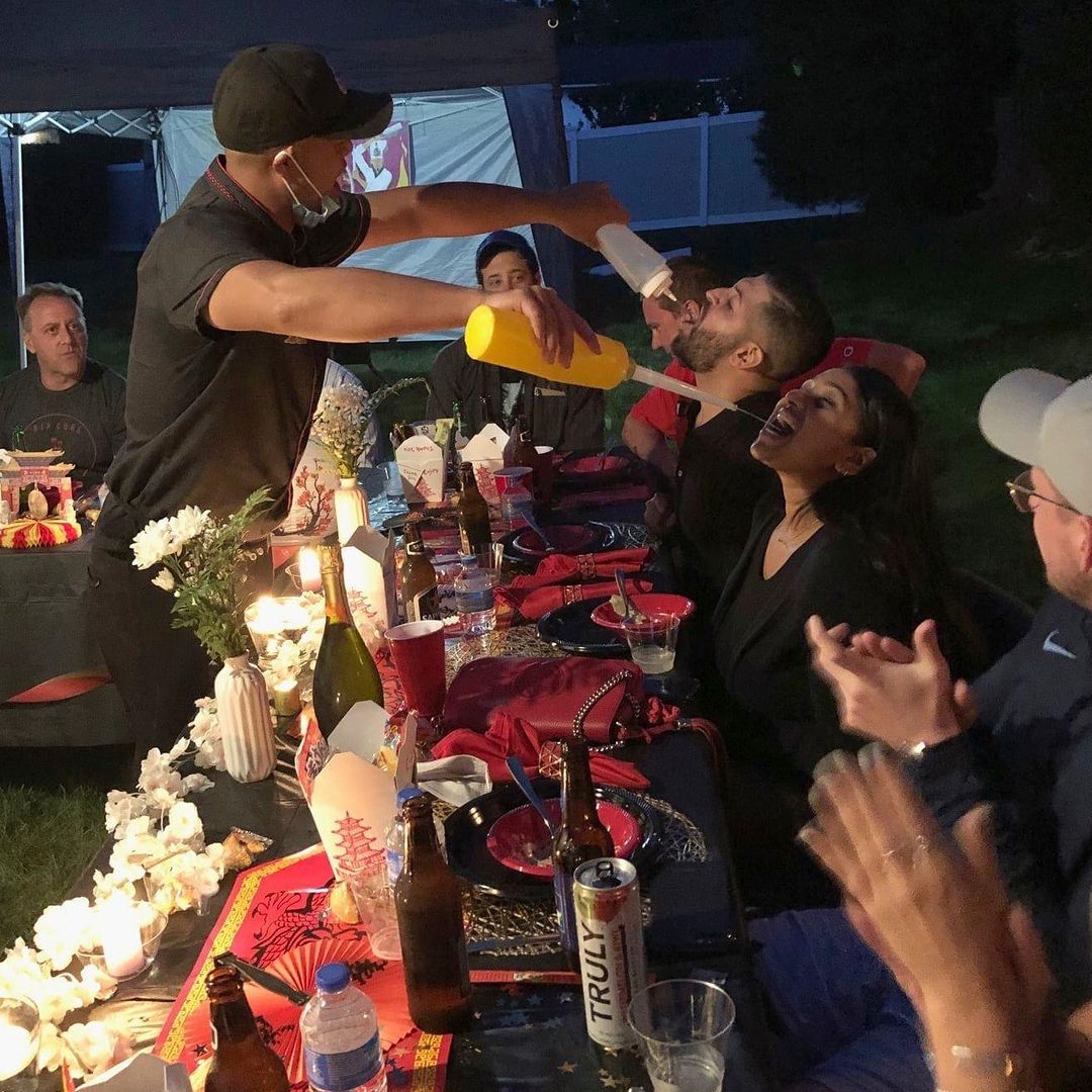 Outdoor evening backyard dinner party at a long table under a canopy; a server theatrically pours from two squeeze bottles into the open mouths of two seated guests while others laugh and clap, table set with takeout boxes, beer bottles, candles and floral decor.