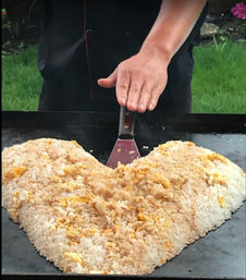 Hand using a spatula to shape heart-shaped fried rice with scrambled egg bits on a flat-top griddle outdoors, casual street-food cooking.