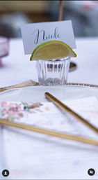 Elegant wedding place setting with a handwritten place card tucked into a small glass topped with a lime wedge, white plate, gold flatware and a floral menu