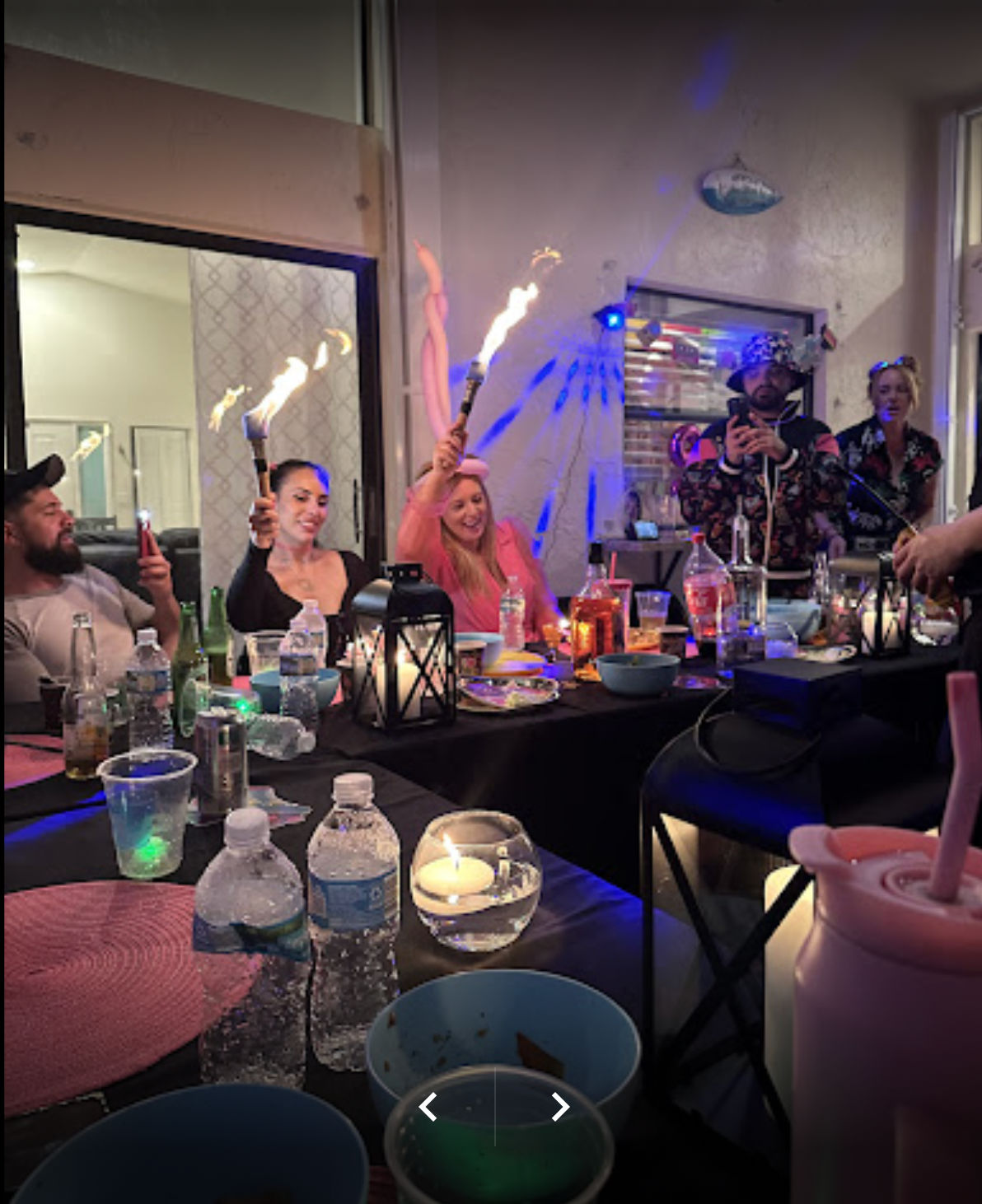 Lively indoor patio party at a long table with drinks, bowls and a lantern; guests cheer while holding flaming sparklers and colorful party lights glow.