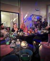 Lively indoor patio party at a long table with drinks, bowls and a lantern; guests cheer while holding flaming sparklers and colorful party lights glow.