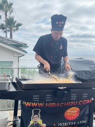 Outdoor hibachi chef cooking fried rice on a flat-top grill at a coastal oceanfront patio with palm trees and a cloudy sky