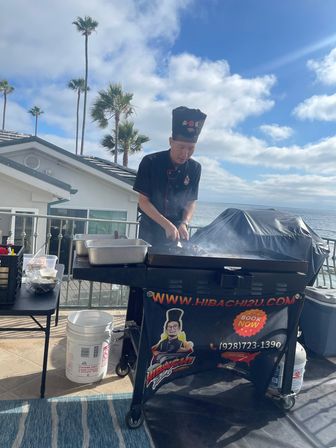 Sizzling hibachi chef cooking on a flat-top grill on a seaside balcony with palm trees and ocean views under a sunny blue sky.