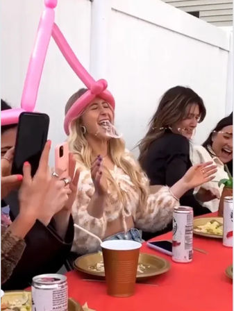 Friends at an outdoor backyard party laughing as a guest wearing a tall pink balloon hat gets a drink or whipped topping splashed on her face; red picnic table with paper plates, cans, cups and phones capturing the moment.