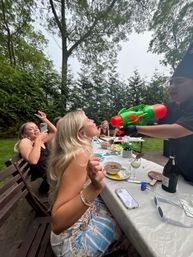 Backyard summer party: woman leaning back laughing as someone squirts water from a bright green-and-red water gun over an outdoor dining table.