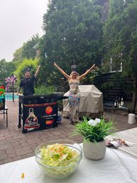 Backyard hibachi-style party on a brick patio — chef at a portable grill and a guest in a chef hat cheering with arms raised, glass bowl of salad and a potted plant on a table in the foreground, pool and lush trees in the background.