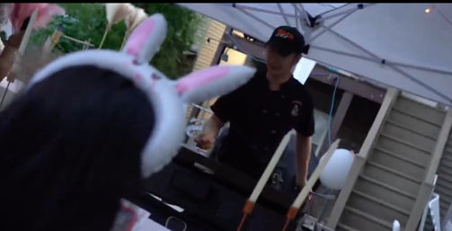 Bunny-eared spectator watches a uniformed food vendor grilling under a white canopy at a backyard outdoor food stall with balloons and a patio fence.