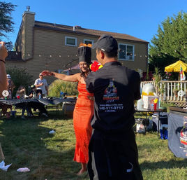 Sunny suburban backyard party with a hibachi chef performing for guests; a woman in a red dress dances wearing a tall flaming chef hat near picnic tables and a house.