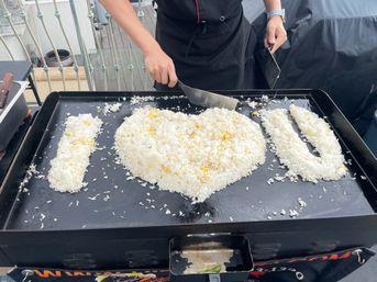 Cook forming playful I LOVE U message from fried rice on a large outdoor flat-top griddle on a rooftop balcony using metal spatulas