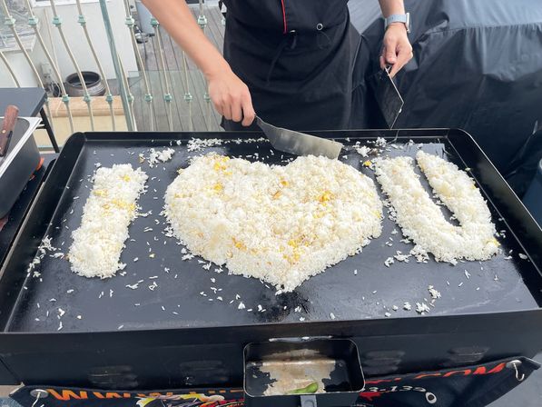Cook forming playful I LOVE U message from fried rice on a large outdoor flat-top griddle on a rooftop balcony using metal spatulas