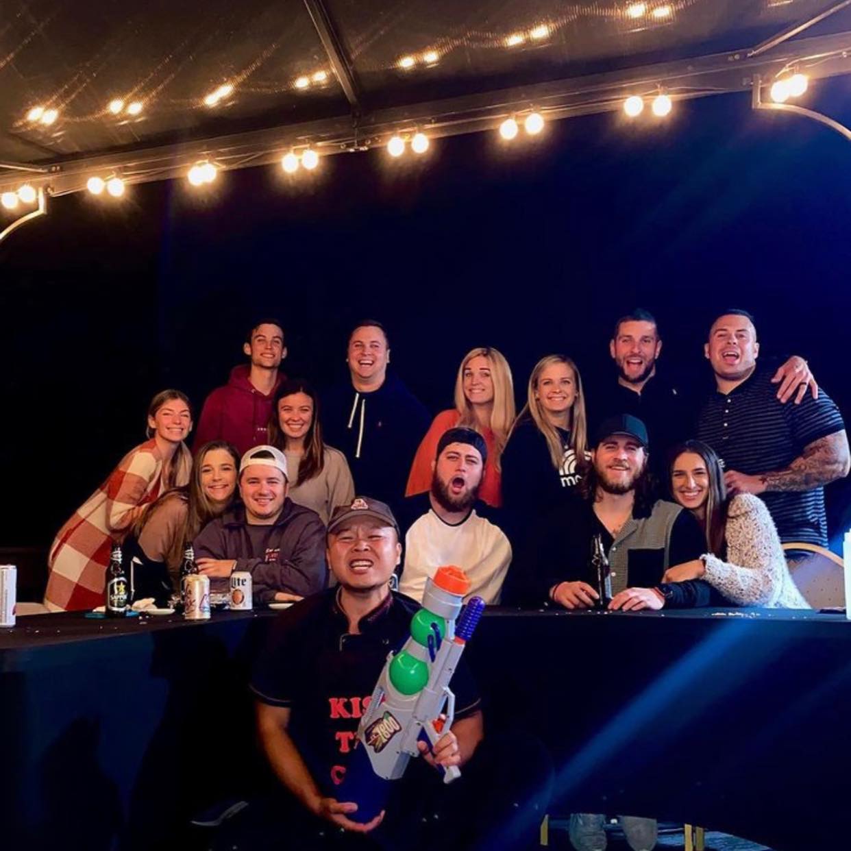 Group photo of friends on a covered outdoor patio at night under string lights, gathered around a table with drinks, one person kneeling in front playfully holding a colorful water gun.