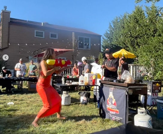 Backyard summer barbecue in a suburban yard: person in a red dress playfully firing a large colorful water gun toward a cook at a grill while guests watch under sunny skies.