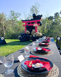 Sunny outdoor garden birthday setup with a long black-clothed table, red-and-black place settings with chopsticks and gold placemats, and a pink torii gate sculpture decorated in black and gold balloons.