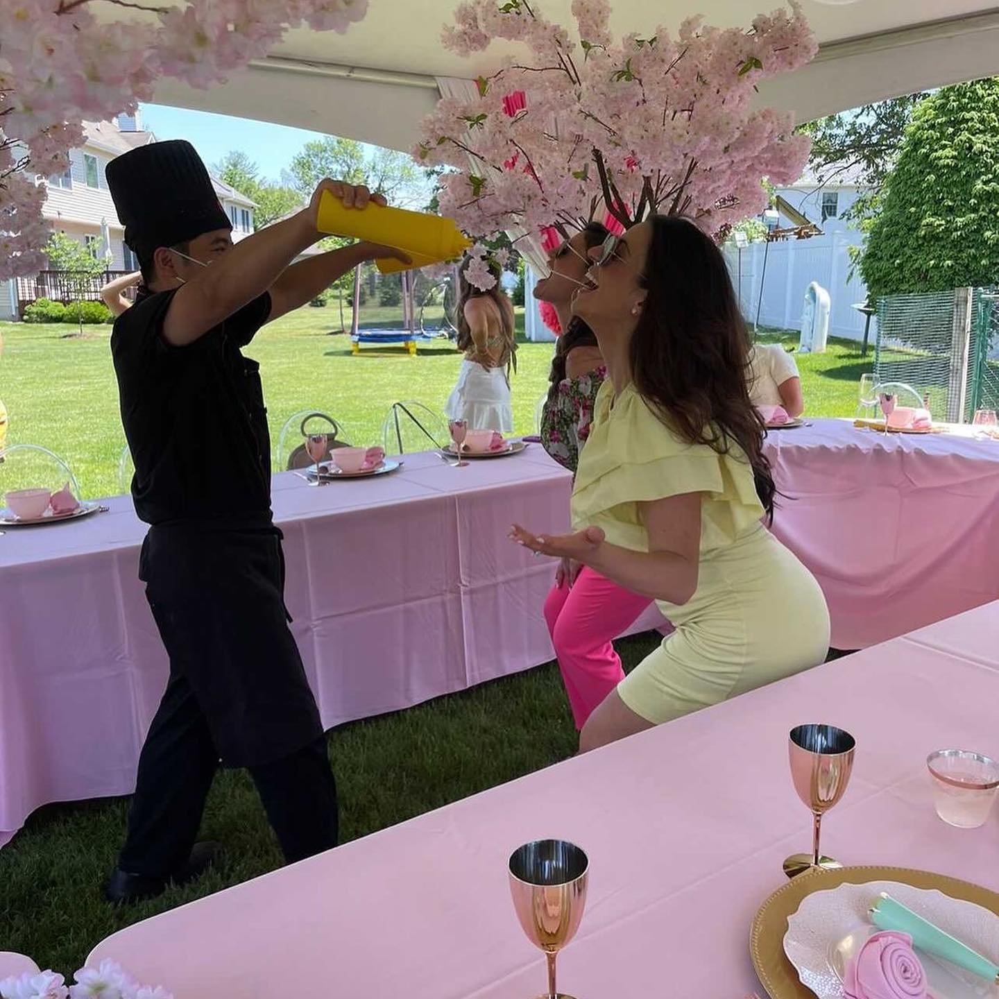 Playful backyard garden party with a chef pouring from a yellow bottle while a woman leans back to catch it beneath a pink cherry-blossom tent; long tables covered in pink tablecloths with tea-party place settings and rose-gold goblets.