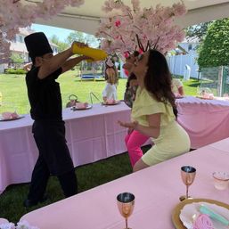 Playful backyard garden party with a chef pouring from a yellow bottle while a woman leans back to catch it beneath a pink cherry-blossom tent; long tables covered in pink tablecloths with tea-party place settings and rose-gold goblets.