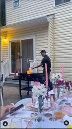 Evening suburban backyard BBQ with a cook flipping food on a flaming outdoor griddle by a beige vinyl-sided house, foreground shows a pink-themed tablescape with taper candles, white daisies and rose-gold glasses.