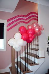 Festive pink-themed indoor staircase with neon-accent wall and white, fuchsia, and pearl balloon garland draped along the wooden banister