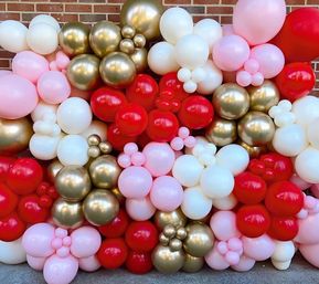 Vibrant balloon garland wall of red, pink, white and metallic gold balloons clustered against a brick wall — festive party photo backdrop
