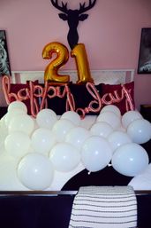 Bedroom party setup with large gold foil "21" balloons and rose-gold "happy birthday" script against a pink wall, white balloons scattered across a bed and black deer head wall decor.