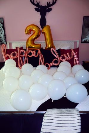 Bedroom party setup with large gold foil "21" balloons and rose-gold "happy birthday" script against a pink wall, white balloons scattered across a bed and black deer head wall decor.