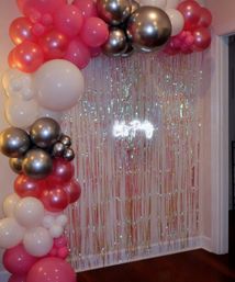 Pink, white and metallic silver balloon garland framing an iridescent fringe curtain with a glowing "Let's Party" neon sign — indoor living-room party backdrop.