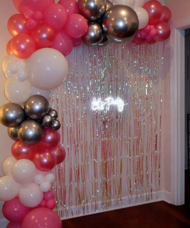 Pink, white and metallic silver balloon garland framing an iridescent fringe curtain with a glowing "Let's Party" neon sign — indoor living-room party backdrop.