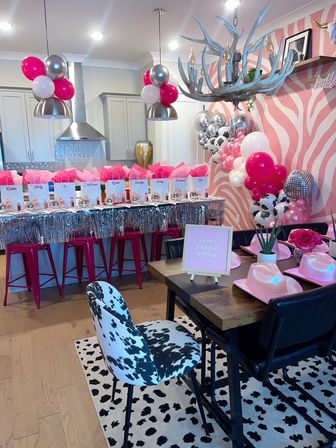 Pink cowgirl-themed birthday party setup in a modern open-plan kitchen and dining area with balloon arches, disco balls, gift bags, pink cowboy hats and zebra-print accents.