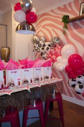 Pink-themed indoor party setup with zebra-print accent wall, metallic and pink balloon garland, disco-ball favors and personalized pink gift bags on a fringe-trimmed table with hot-pink stools — festive birthday party decorations.