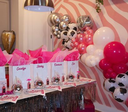Pink-themed indoor party setup with zebra-print accent wall, metallic and pink balloon garland, disco-ball favors and personalized pink gift bags on a fringe-trimmed table with hot-pink stools — festive birthday party decorations.