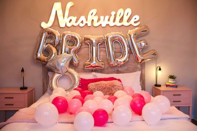 Cozy bedroom styled for a bachelorette: illuminated 'Nashville' sign and silver 'BRIDE' and diamond ring balloons above a bed strewn with pink and white balloons, flanked by bedside tables with lamps and a small plant.