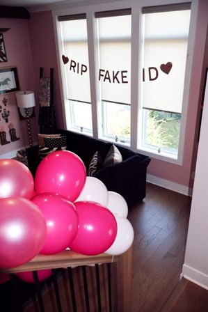 Pink and white party balloons clustered on a stair railing in a pink-walled home living room, featuring a black couch with patterned pillows and a glittery "RIP FAKE ID" banner strung across three tall windows.