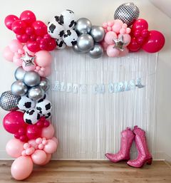 Pink, hot-pink and silver balloon garland with cow-print and mirrored disco balloons framing a metallic fringe backdrop, glitter "Let’s Go Girls" banner and pink cowboy boot props — cowgirl party photo backdrop