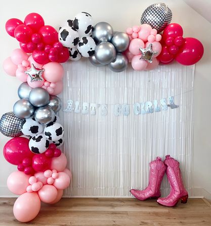 Pink, hot-pink and silver balloon garland with cow-print and mirrored disco balloons framing a metallic fringe backdrop, glitter "Let’s Go Girls" banner and pink cowboy boot props — cowgirl party photo backdrop