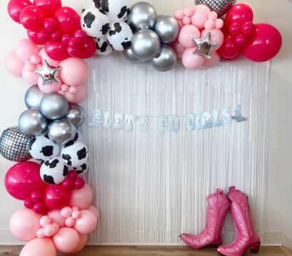 Pink, hot-pink and silver balloon garland with cow-print and mirrored disco balloons framing a metallic fringe backdrop, glitter "Let’s Go Girls" banner and pink cowboy boot props — cowgirl party photo backdrop