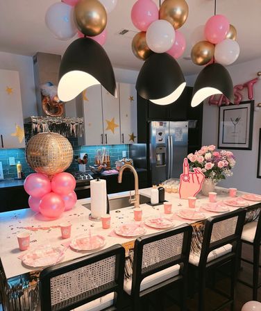 Festive modern kitchen island decorated for a party with pink, white and gold balloons, a gold disco-ball column, black pendant lights, pink plates and cups, and a bouquet of pink roses