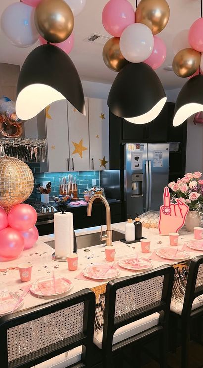 Festive modern kitchen island decorated for a party with pink, white and gold balloons, a gold disco-ball column, black pendant lights, pink plates and cups, and a bouquet of pink roses