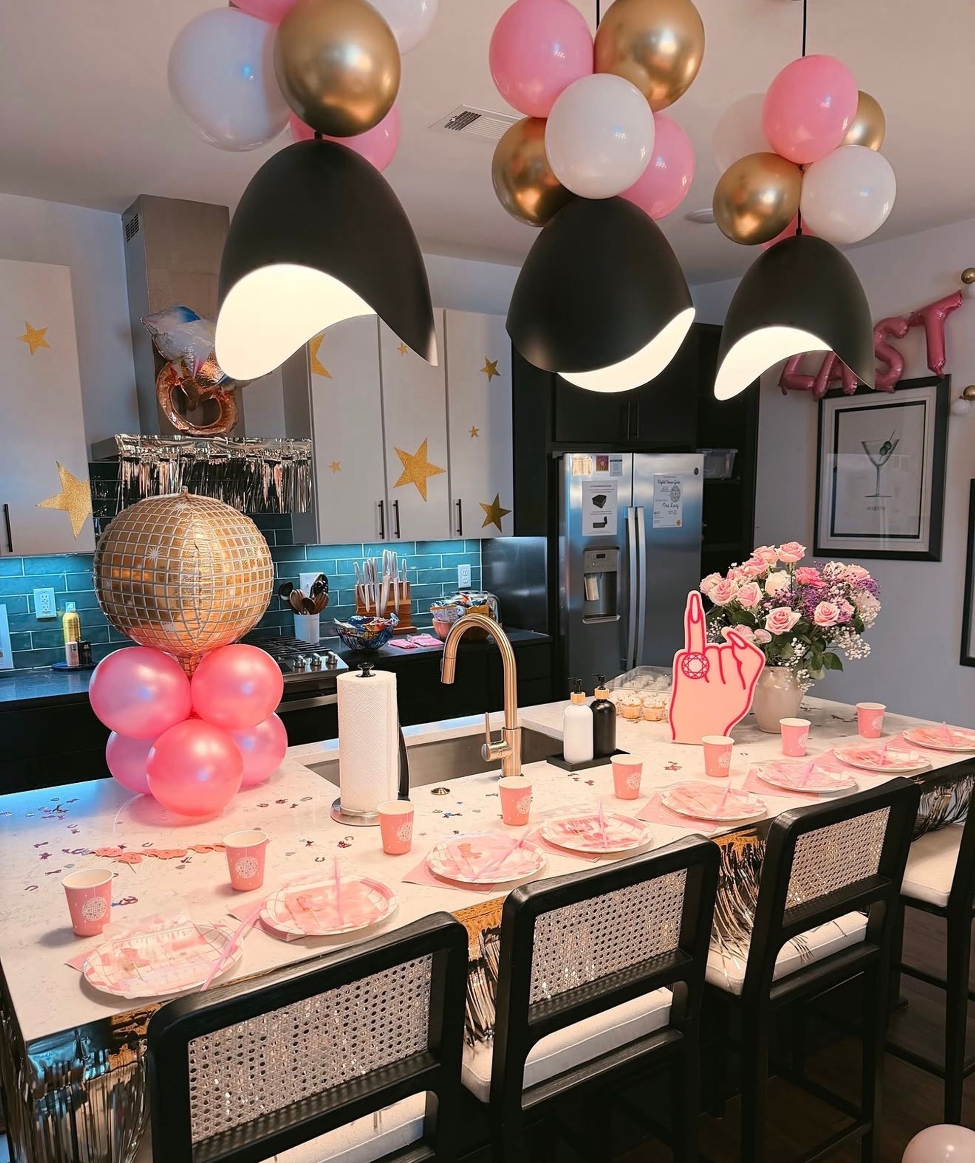 Festive modern kitchen island decorated for a party with pink, white and gold balloons, a gold disco-ball column, black pendant lights, pink plates and cups, and a bouquet of pink roses