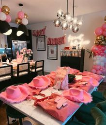 Pink rodeo-themed bachelorette party setup in a modern dining/kitchen area — marble table with pink feathered cowboy hats, matching tote bags and sashes, balloon garlands, floral centerpiece, metallic pendant lights and a neon "this must be the place" sign.