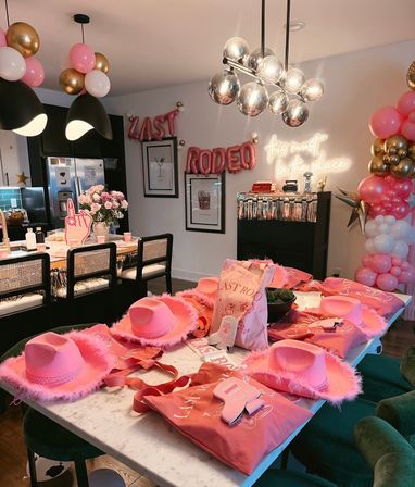 Pink rodeo-themed bachelorette party setup in a modern dining/kitchen area — marble table with pink feathered cowboy hats, matching tote bags and sashes, balloon garlands, floral centerpiece, metallic pendant lights and a neon "this must be the place" sign.