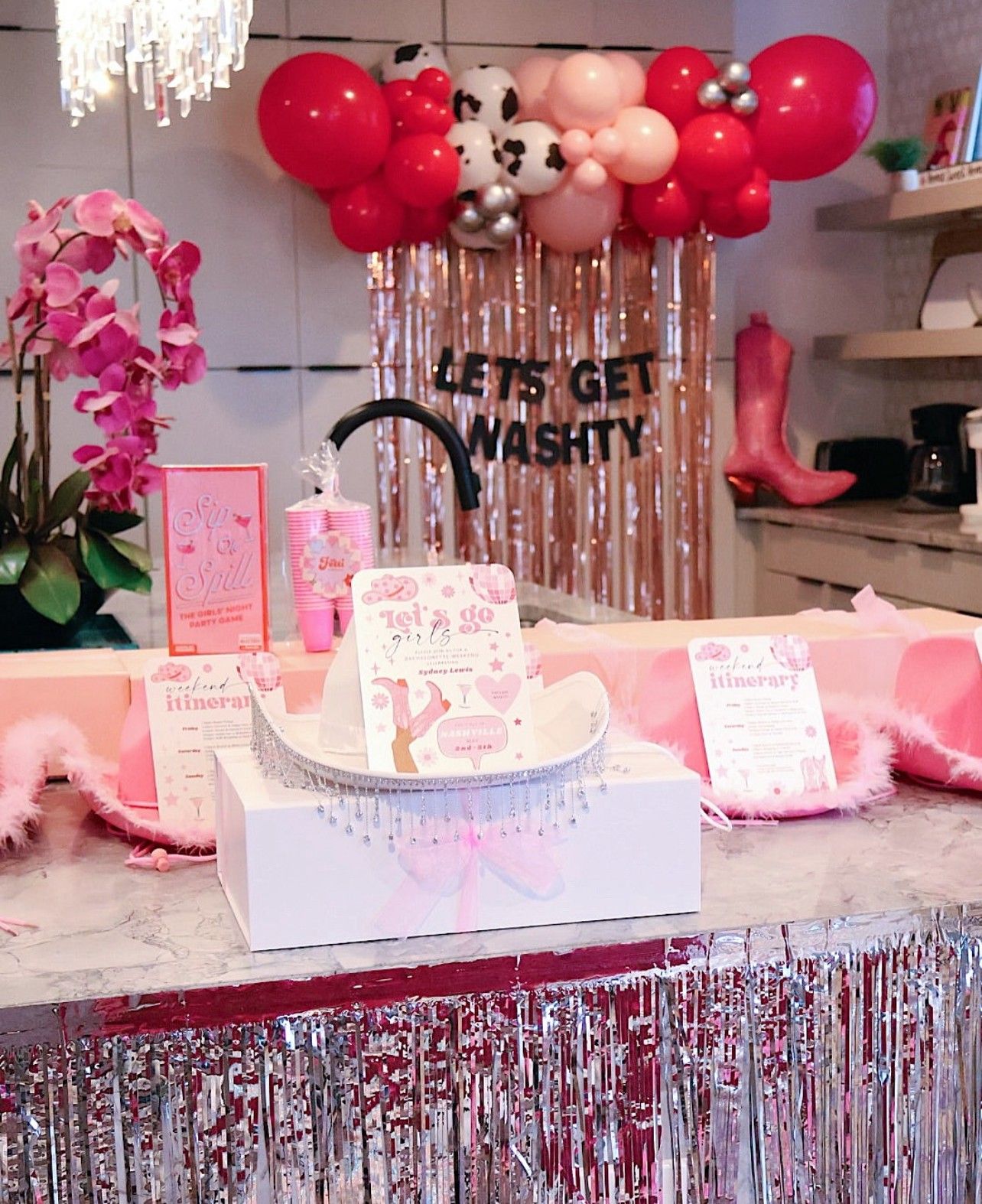 Pink cowgirl-themed bachelorette party setup on a kitchen island with red and pink balloon garland, metallic fringe backdrop reading "LET'S GET WASHTY", pink cowboy boot prop, orchid, feather boas, tiara and itinerary cards — playful pink party decor.