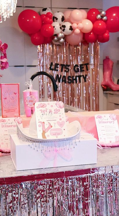 Pink cowgirl-themed bachelorette party setup on a kitchen island with red and pink balloon garland, metallic fringe backdrop reading "LET'S GET WASHTY", pink cowboy boot prop, orchid, feather boas, tiara and itinerary cards — playful pink party decor.