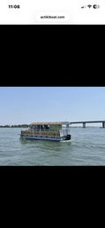 Tiki-themed pontoon boat with a thatched roof carrying passengers on calm coastal waters, cruising past a low concrete bridge under a clear blue sky.