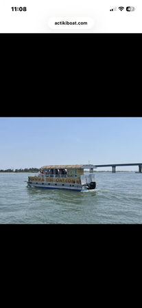 Tiki-themed pontoon boat with a thatched roof carrying passengers on calm coastal waters, cruising past a low concrete bridge under a clear blue sky.