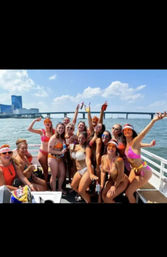 Boat party of a dozen women in colorful bikinis and visors, smiling and holding drinks on a sunny day with a city skyline and bridge beyond calm harbor waters